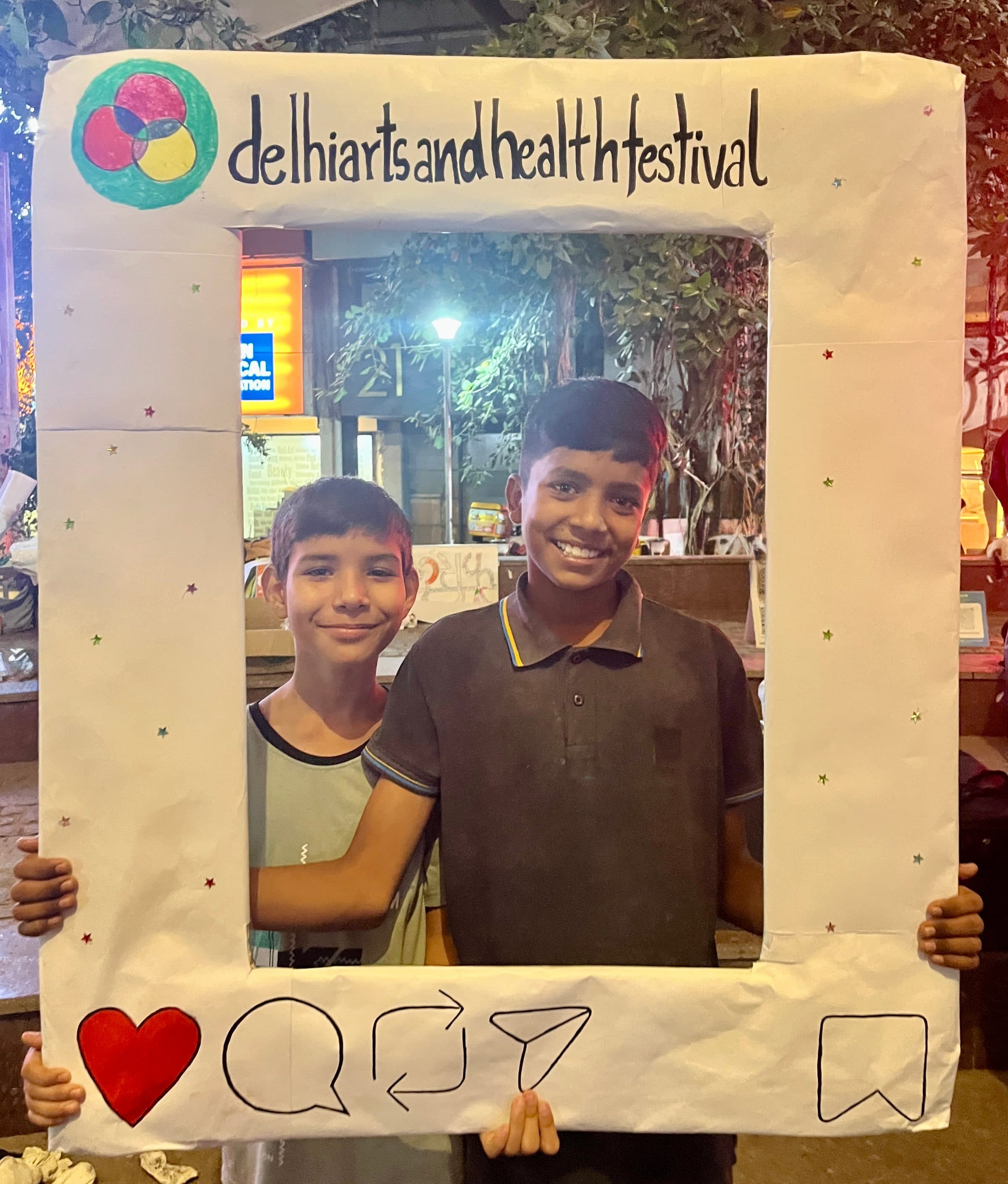 Two children posing together, smiling, inside a hand-decorated Delhi Arts and Health Festival photo frame