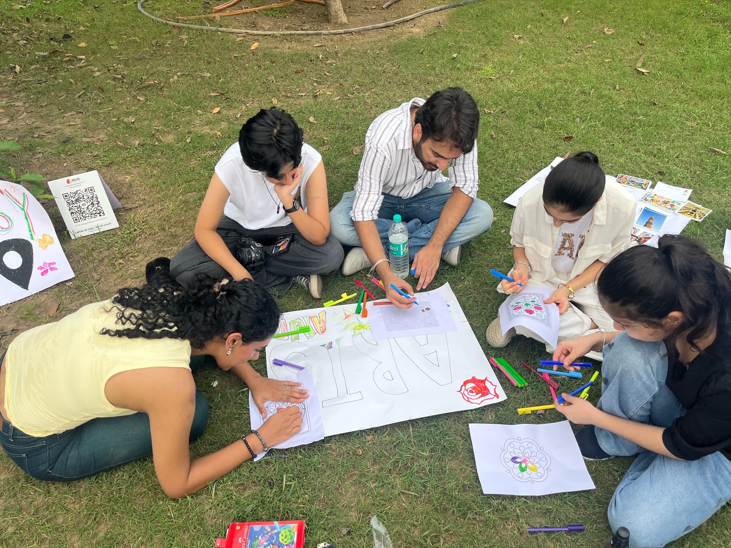 Five people sitting on the grass around a large hand-drawn ARTS poster, colouring with markers